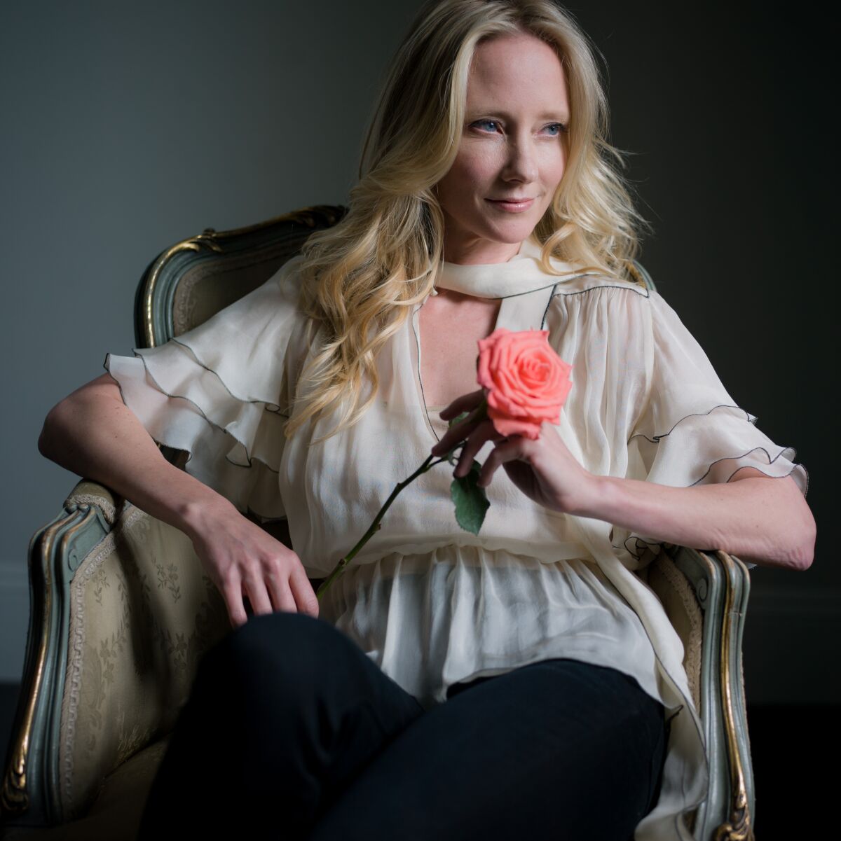 Anne Heche sits in a chair, looking left, and holding a pink rose