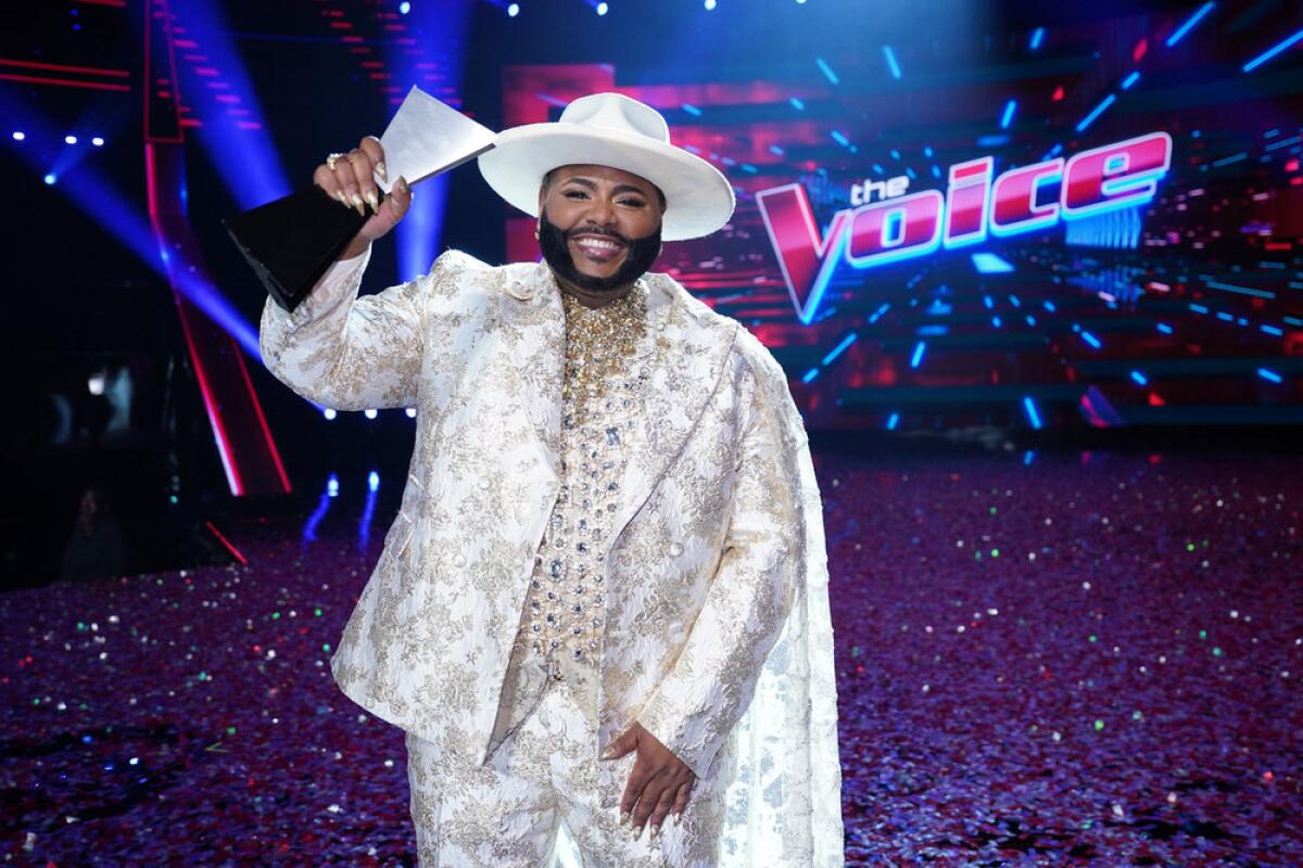Asher HaVon, wearing a white suit and hat, holds up a trophy in front of "The Voice" logo.