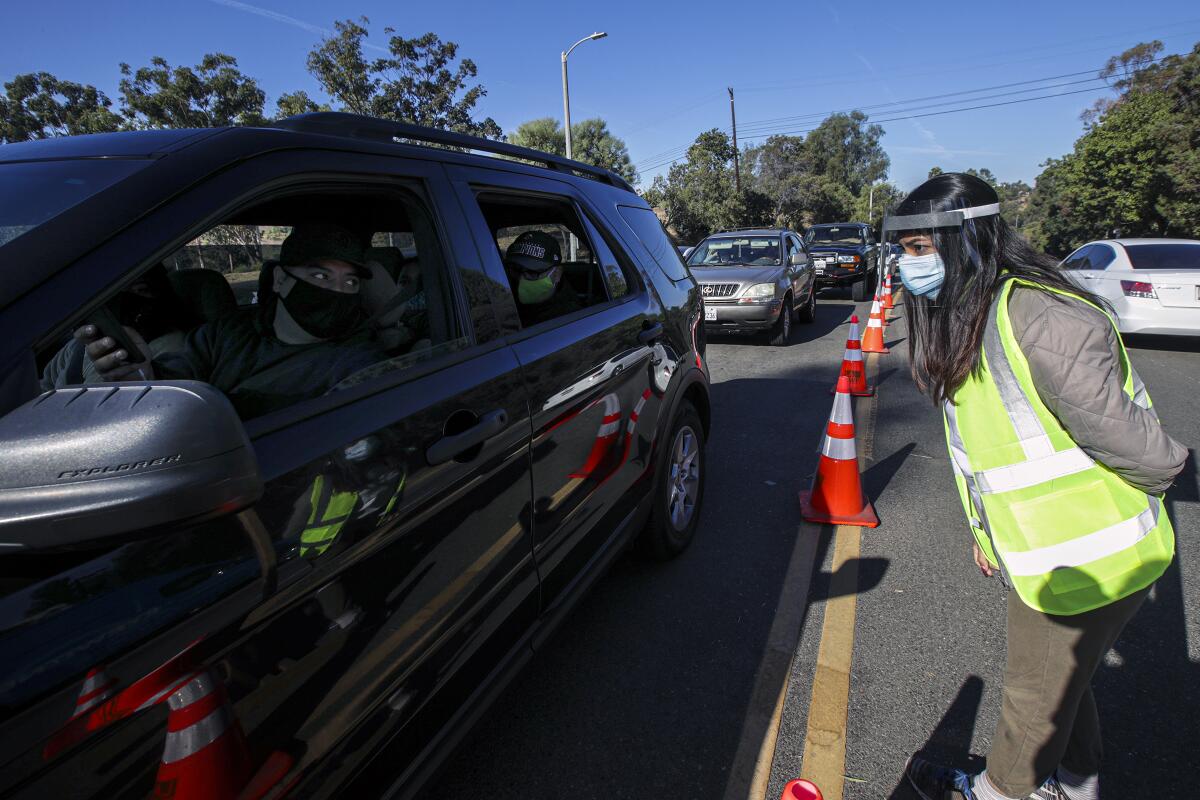 A woman in a yellow safety vest with a face shield speaks to a masked driver in a line of waiting cars.