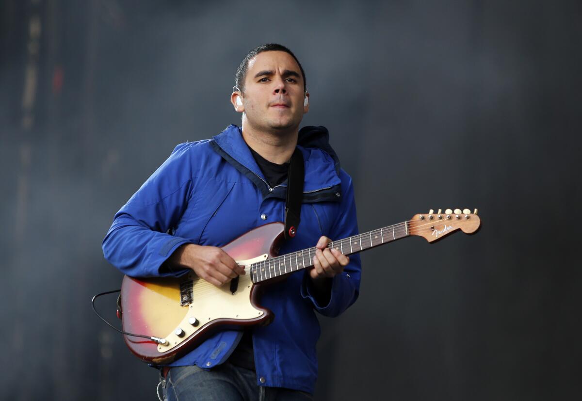 Rostam Batmanglij, wearing a blue jacket, plays the electric guitar.