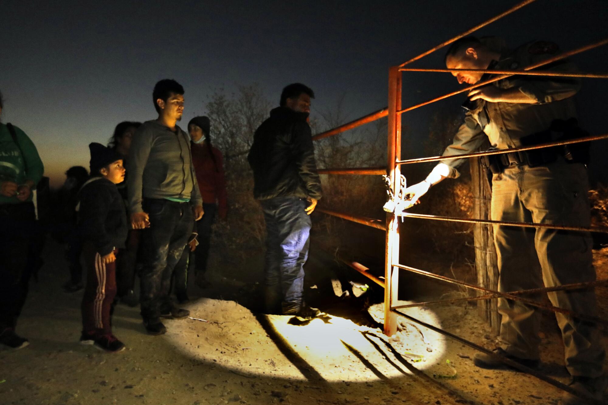 Migrants seeking asylum wait by a fence as a Border Patrol agent shines his flashlight toward them.