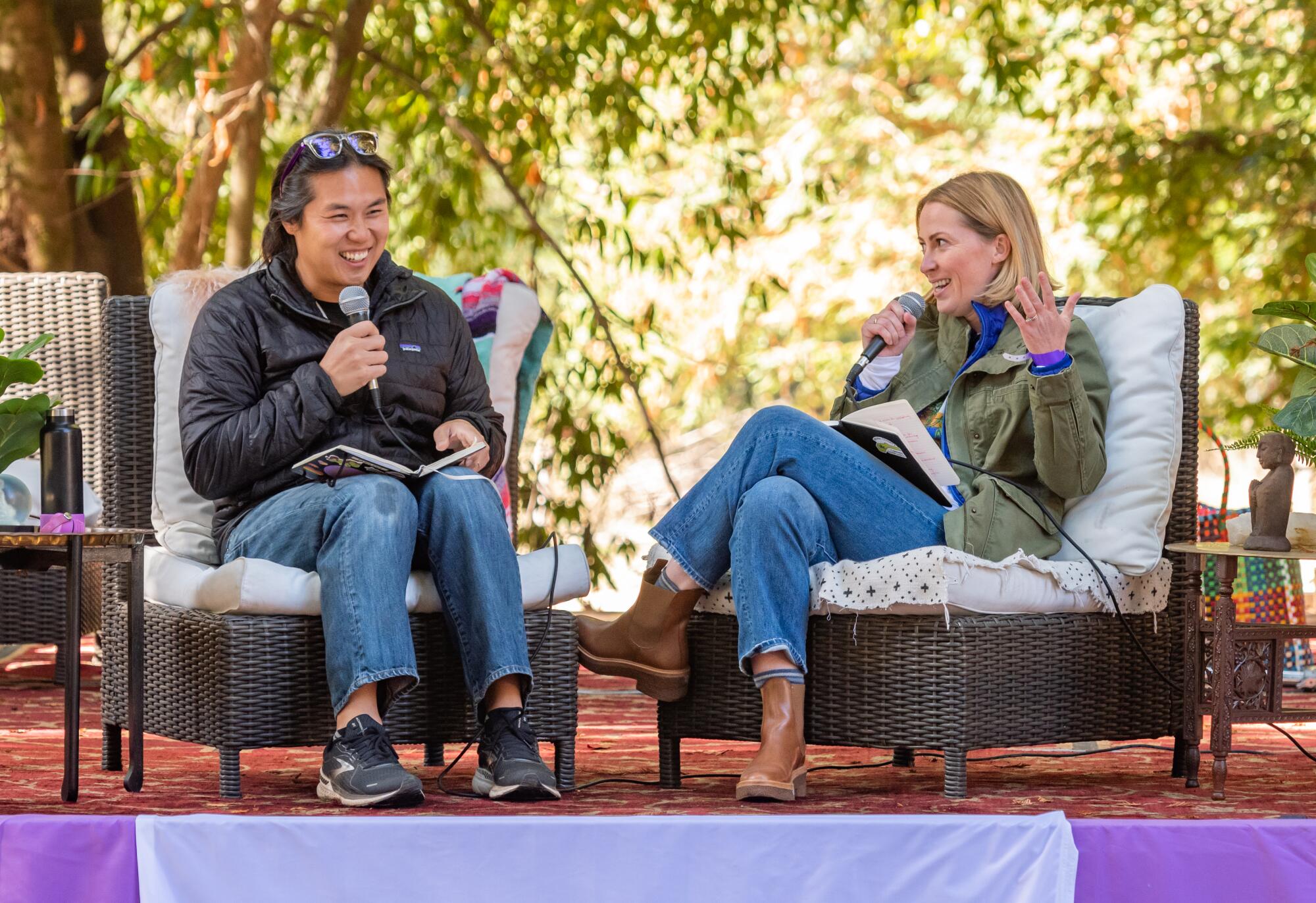 A man, left, and a woman hold microphones as they sit and talk on an outdoor stage