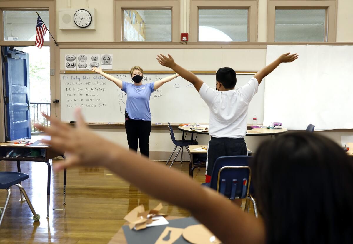 A masked teacher leads children in a stretching exercise in a classroom.