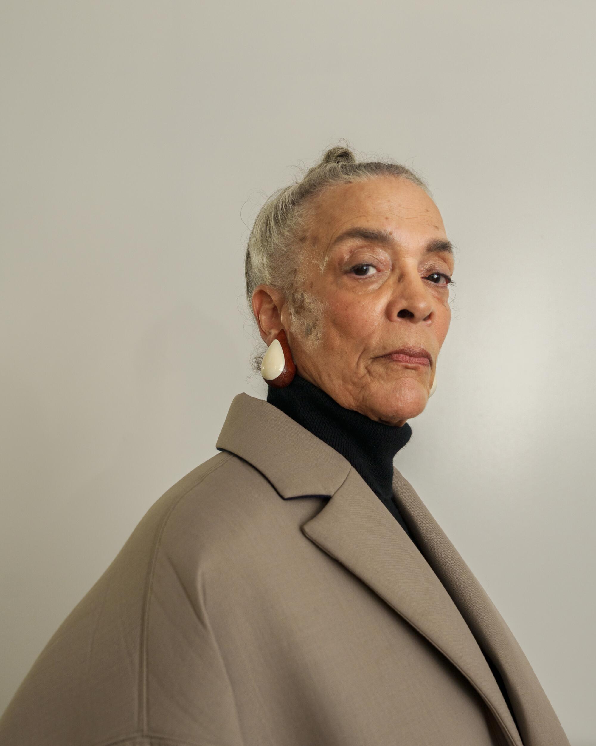 A photo of a woman wearing white earrings, a black turtle neck, and a brown jacket.