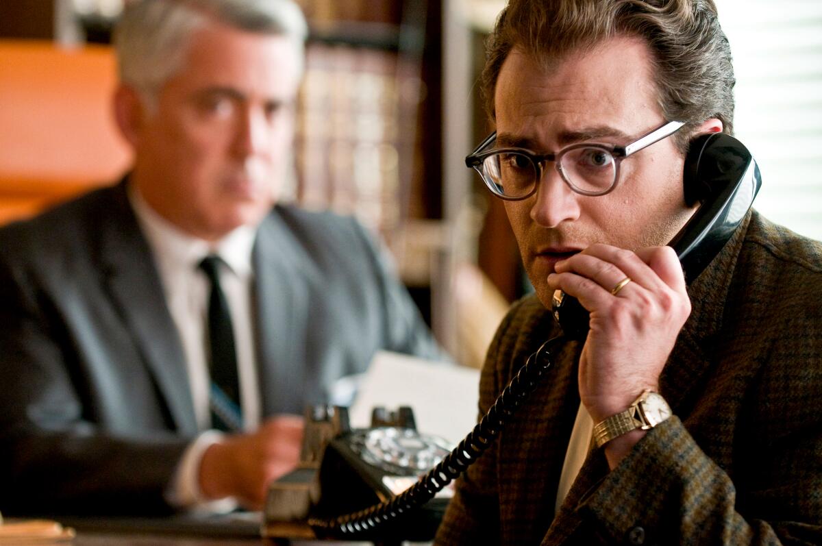 A man talks on a landline phone looking concerned while another man in a suit sits at a desk behind him in "A Serious Man." 