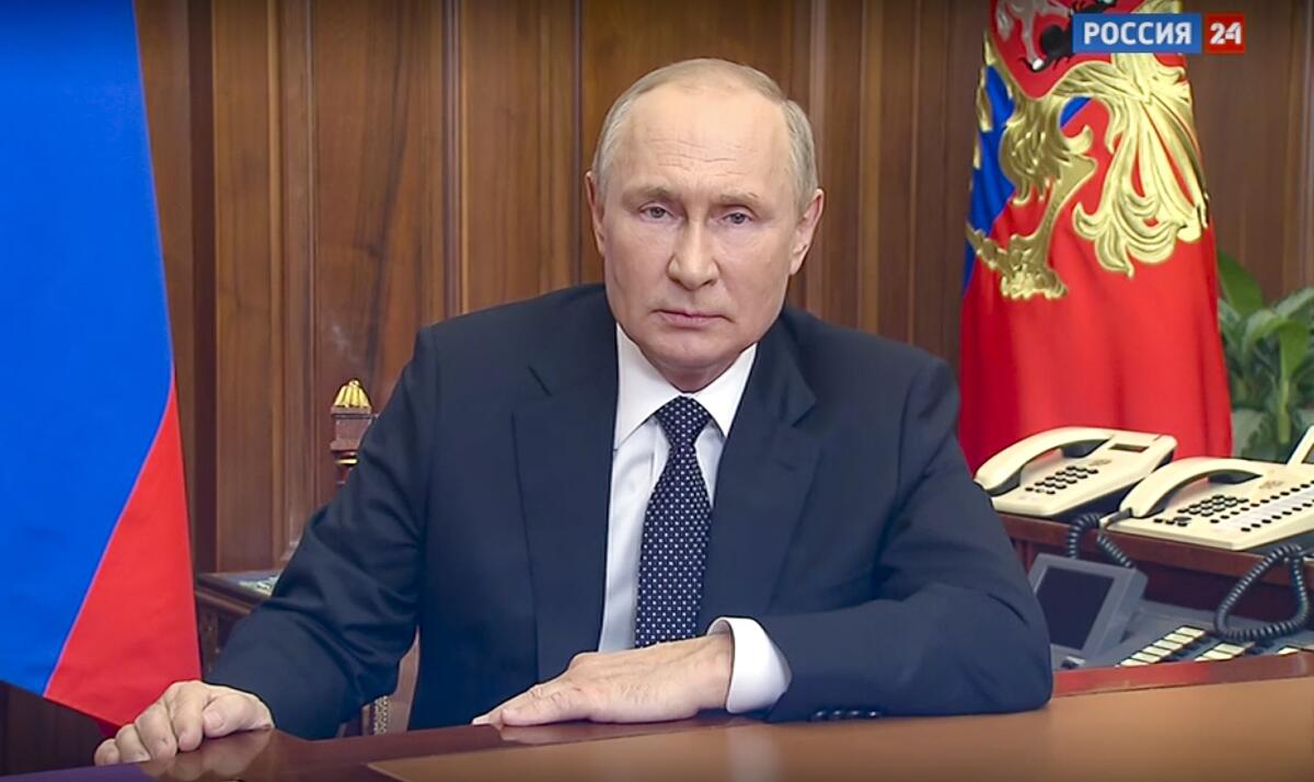 Vladimir Putin sits at a desk during a TV broadcast.