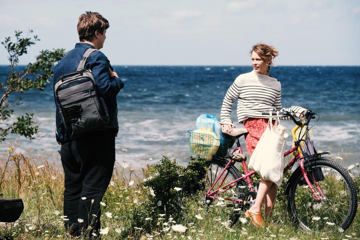 A man with a backpack speaks to a woman on a bicycle by the seashore