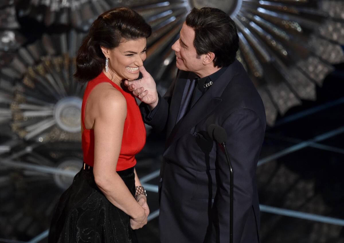 John Travolta cups Idina Menzel's chin as they face each other on a stage.