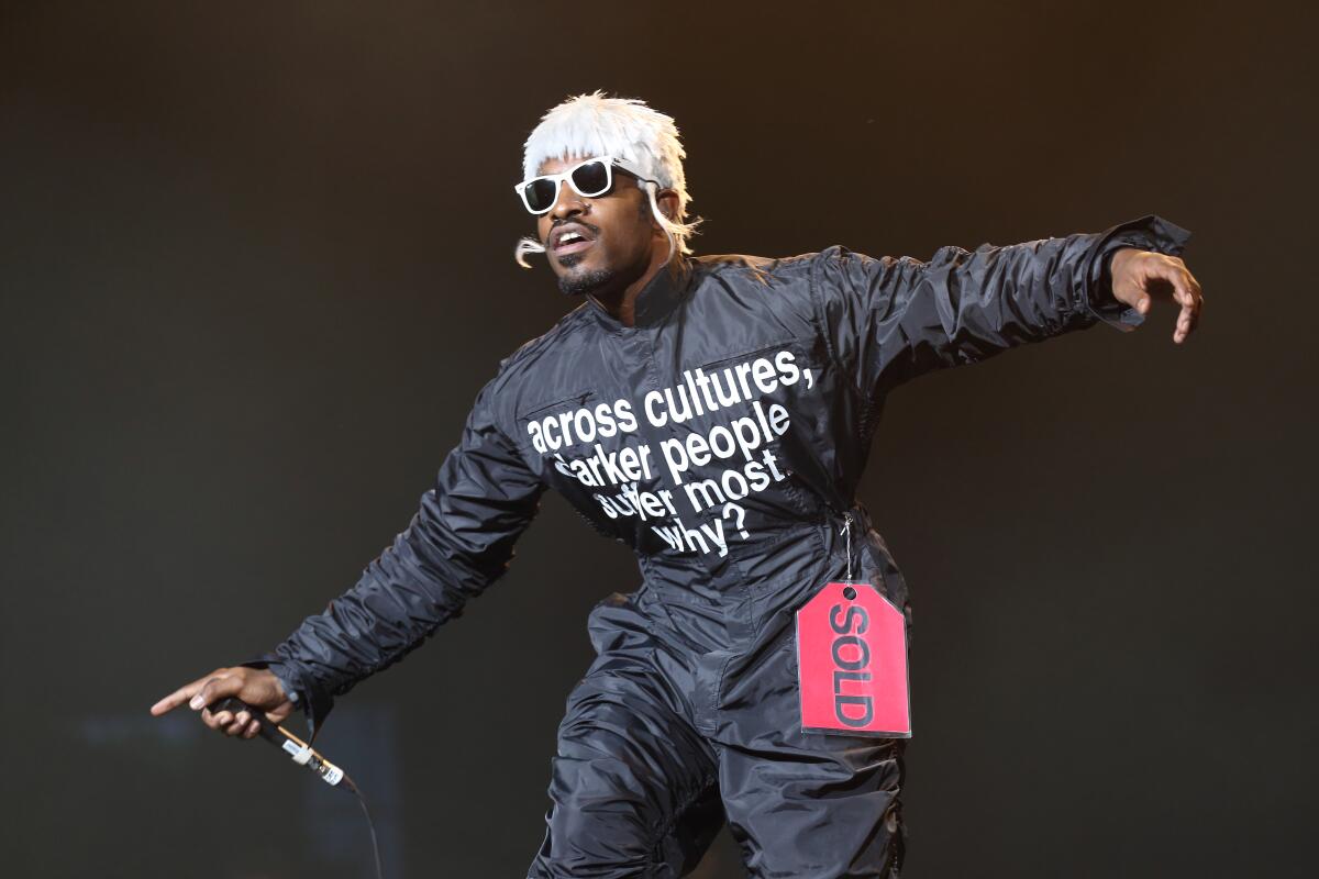 Andre 3000 in a black jumpsuit, short white wig and sunglasses holding a microphone on a stage