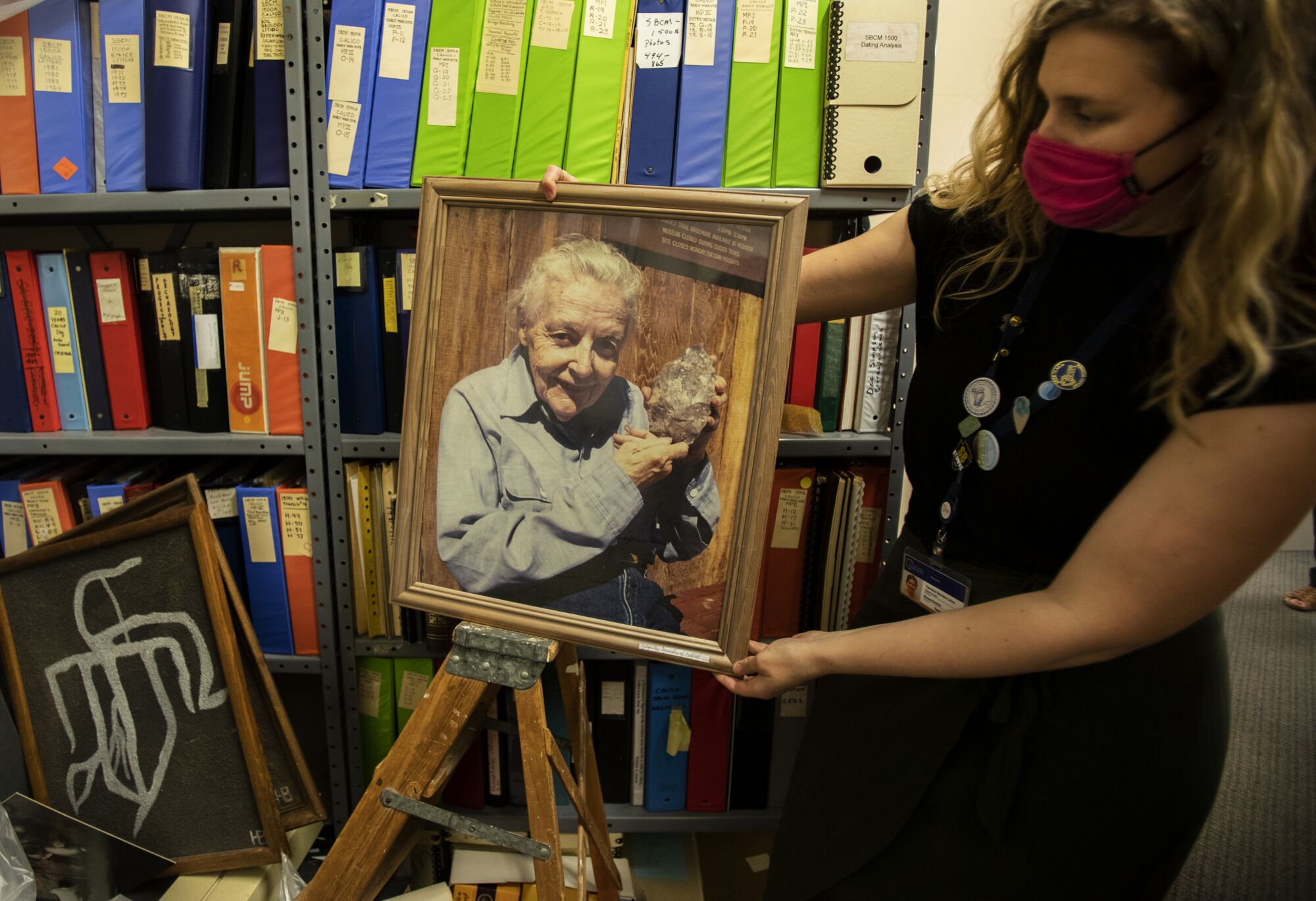 A woman displays a framed portrait of an older woman holding up an ancient stone tool.