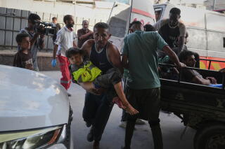 A man rushes forward while carrying a child next to people in the back of a truck and a van with its rear doors open