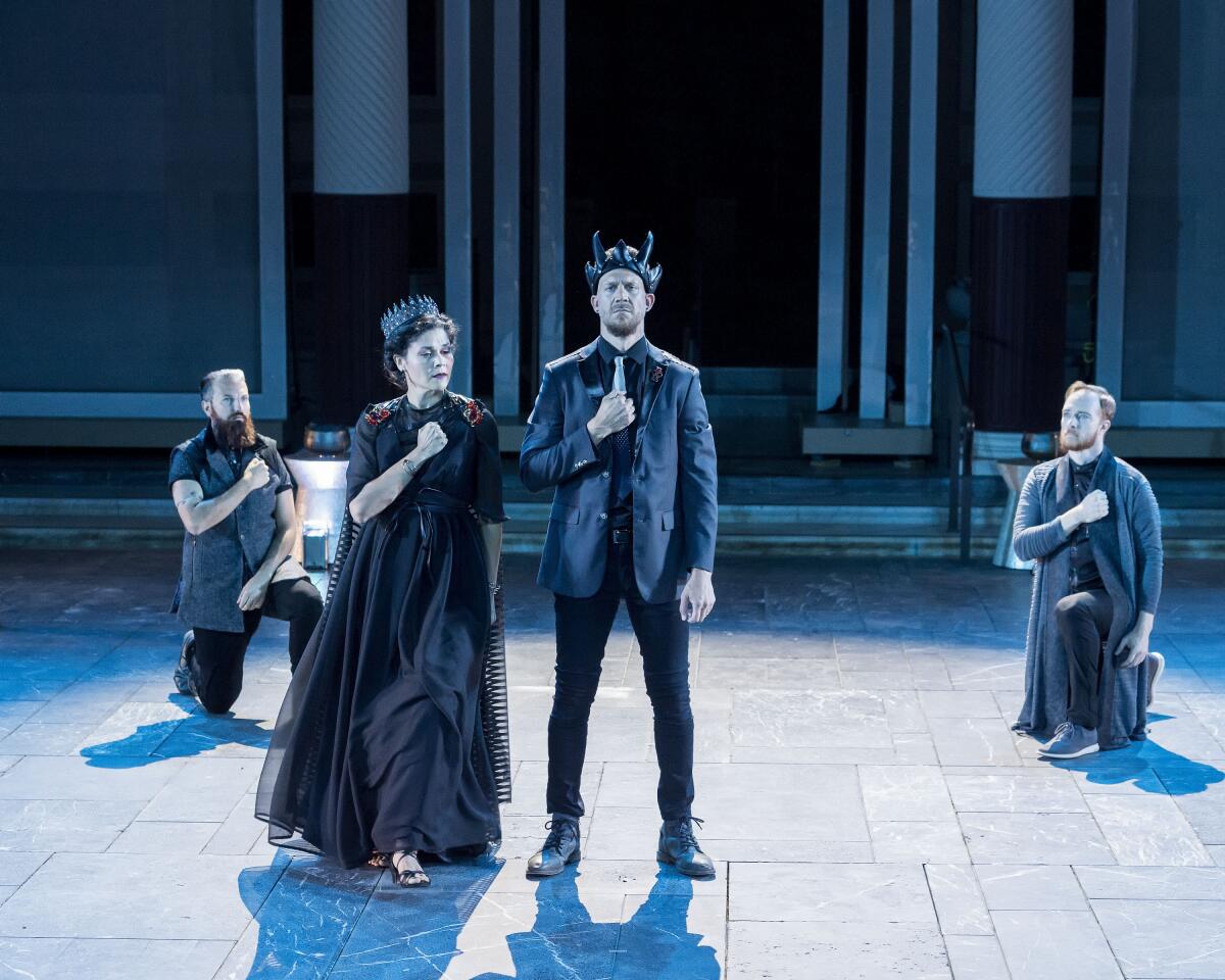 A woman in a long gown and cape and a man in a crown onstage. Behind them two men kneel.