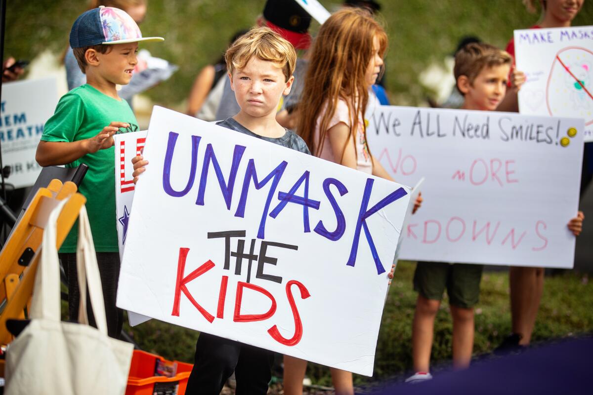 A child at a protest holds a sign reading "Unmask the Kids."