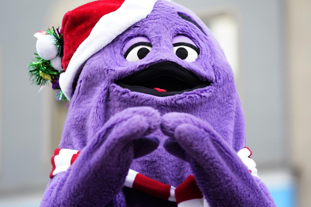 A furry purple creature holds its hands below its face while wearing a Santa hat and a red and white scarf