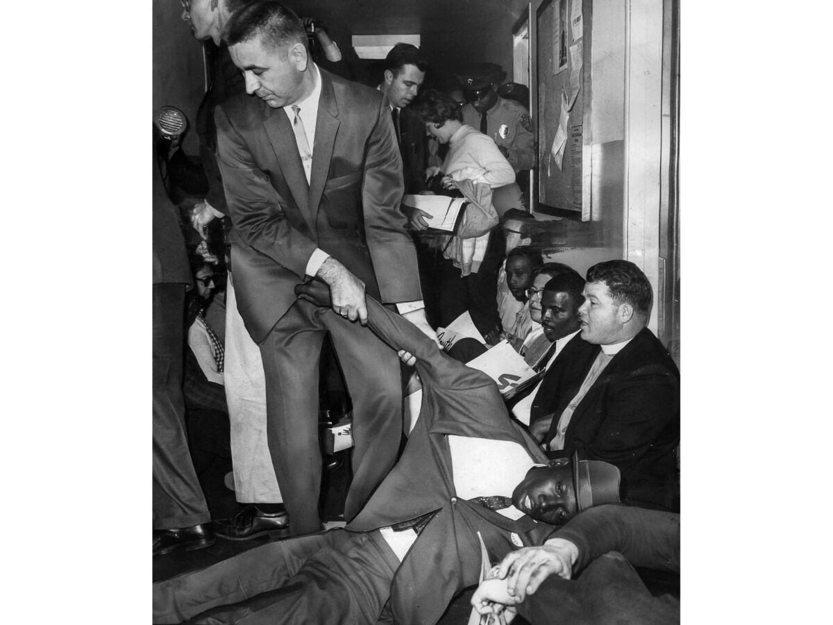 A white man, standing, drags a prone Black demonstrator, who appears limp, by the arm, amid a row of sit-in demonstrators.