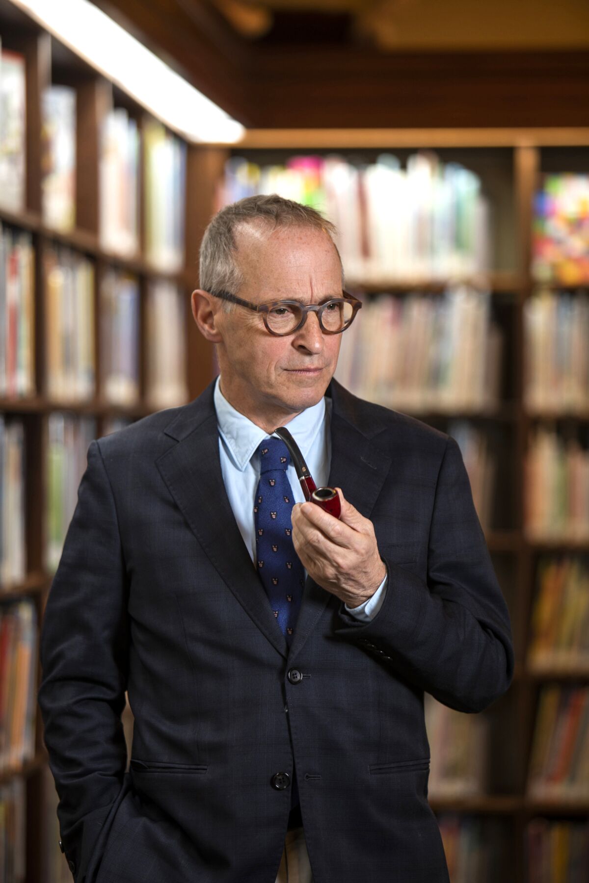 A man, wearing a suit and tie and holding a pipe, stands among bookshelves.