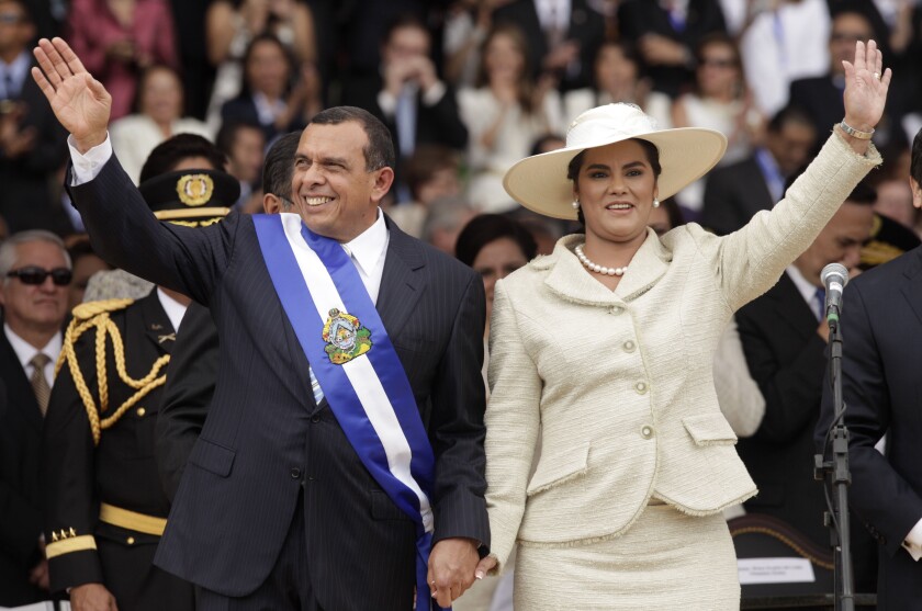 A man in a suit and blue and white sash and a woman in a white suit and hat wave as they hold hands 