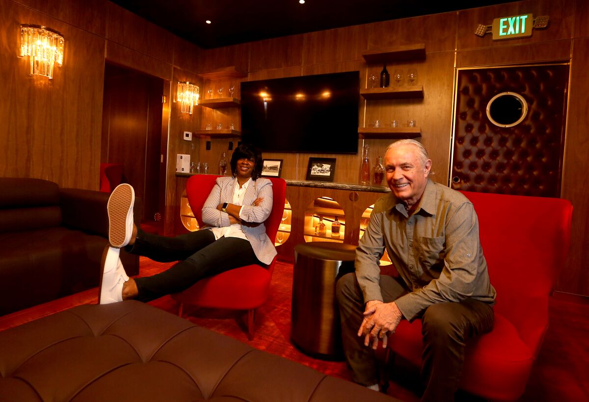 Man and woman sit in red lounge chairs in a wood paneled room