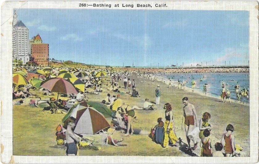 Colorful umbrellas and swimsuits dot an expanse of sand and shore, with a jetty and tall buildings in the background