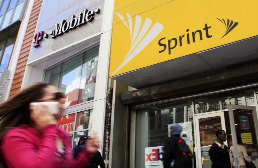 A woman holding her phone to her ear walks past adjacent T-Mobile and Sprint stores.
