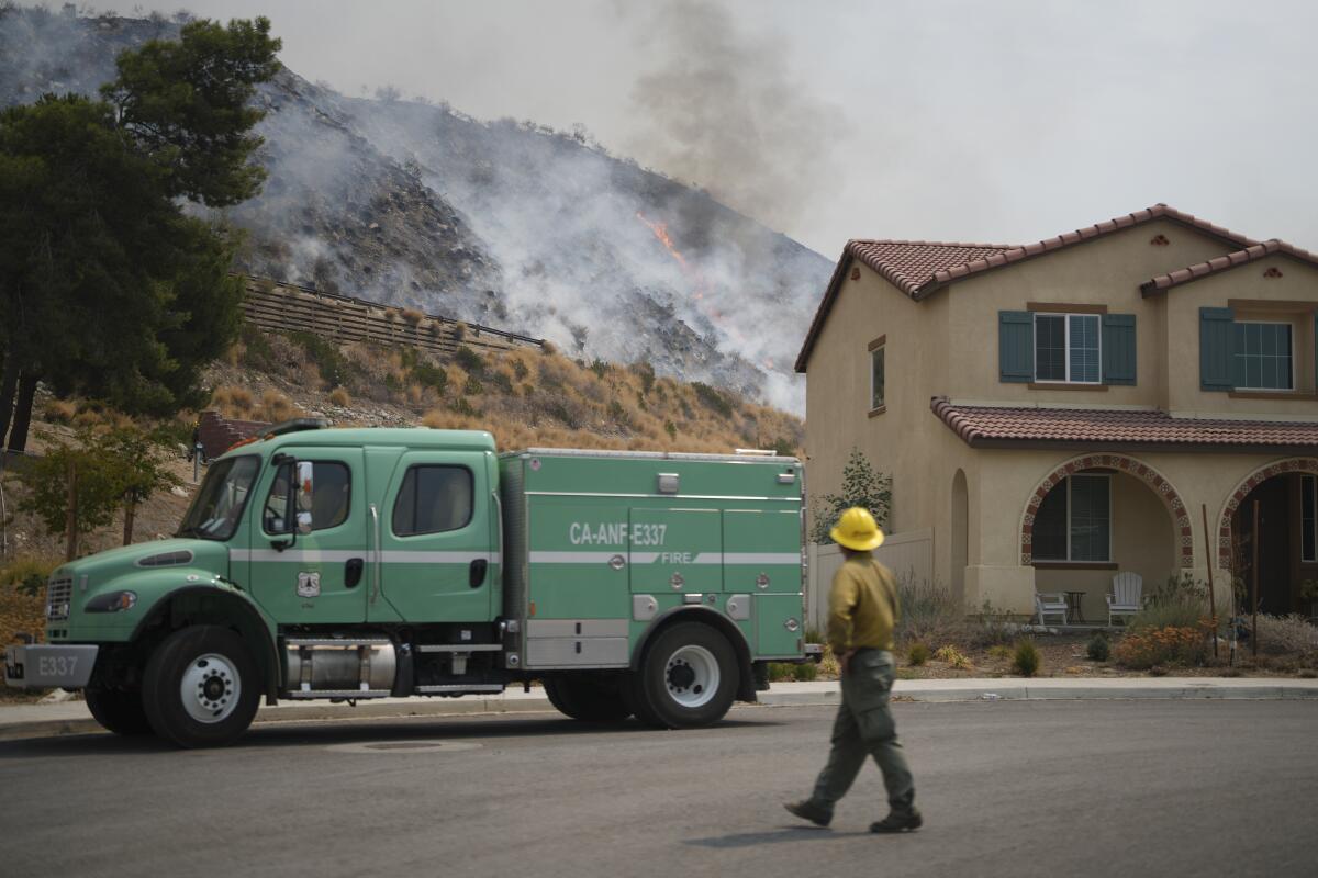 A person walks in front of a truck and a house with a forest fire in the background.