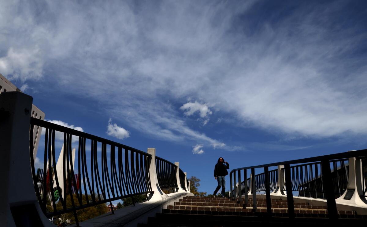A woman crosses the pedestrian bridge over Temple Street against a cloud-filled sky in downtown Los Angeles.
