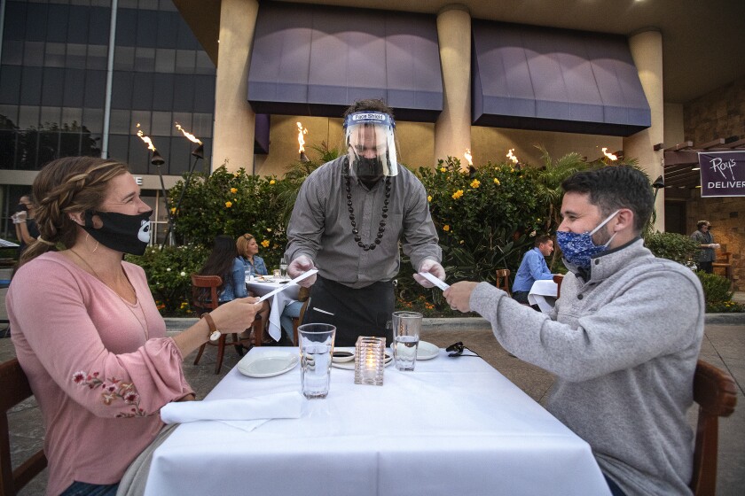 A man wearing a mask and plastic face shield hands chopsticks to diners seated outdoors at Roy's restaurant