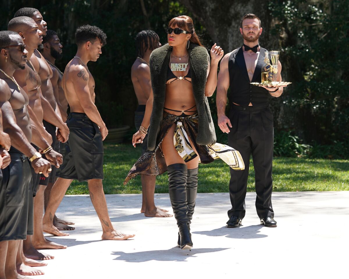 A woman in a bikini, thigh-high boots and fur vest walks in front of a line of shirtless men.