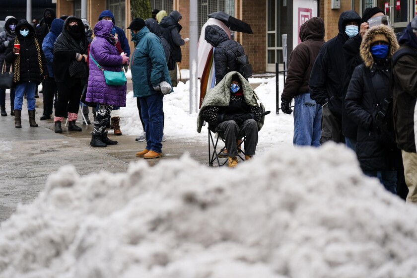 People in winter coats and blankets stand, or sit, in a long line amid snow.