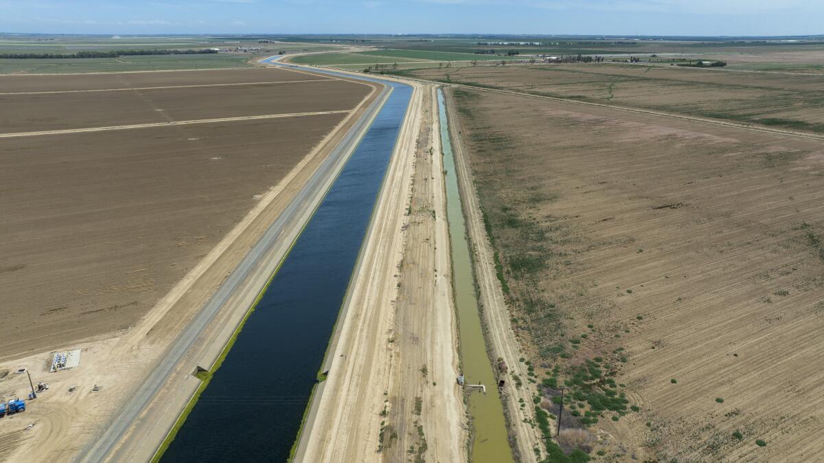 A large canal and smaller irrigation channel run parallel to one another across dusty farmland.
