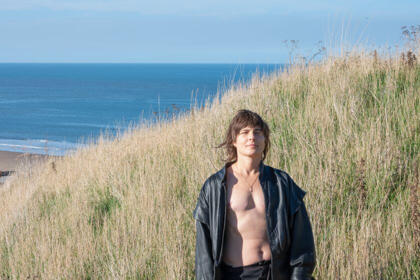 A nonbinary trans man stands in a field of sea grass in an open sweatshirt