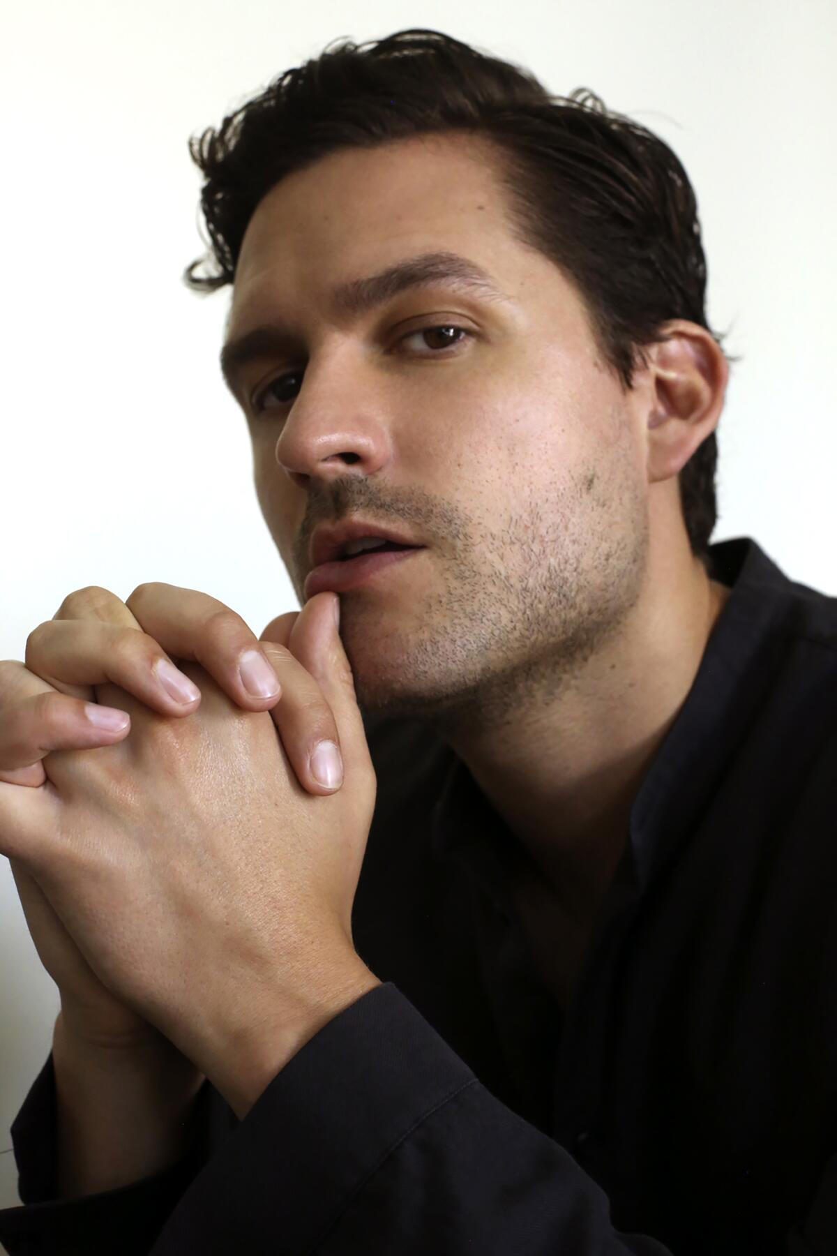 A close-up portrait of a man, his hands clasped in front of his chin