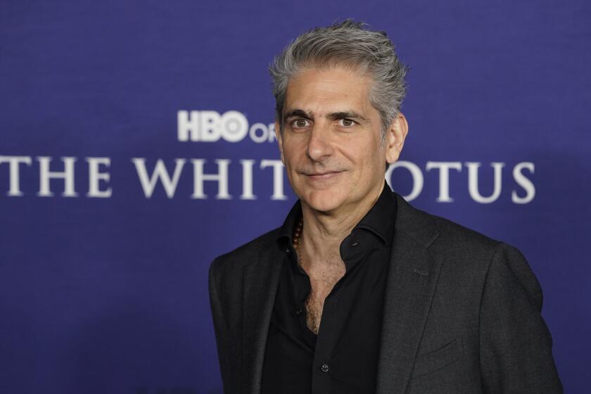 Michael Imperioli posing in a gray suit and black shirt against a purple background.