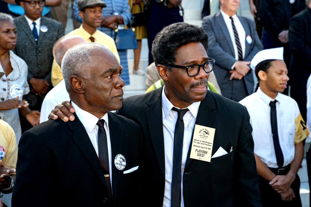 Two men in suits stand amid a March on Washington crowd in a scene from "Rustin."