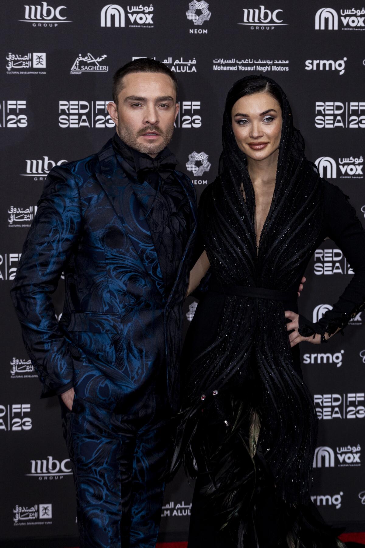 Ed Westwick and Amy Jackson dressed in dark formalwear pose together for a photo in front of a backdrop