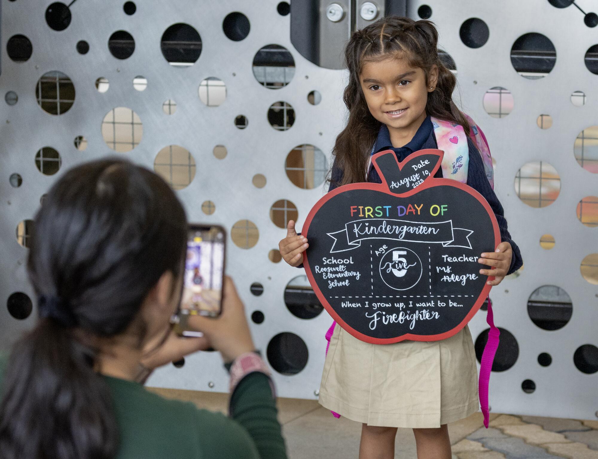 A child holds an apple-shaped chalkboard that reads, "First day of kindergarten."