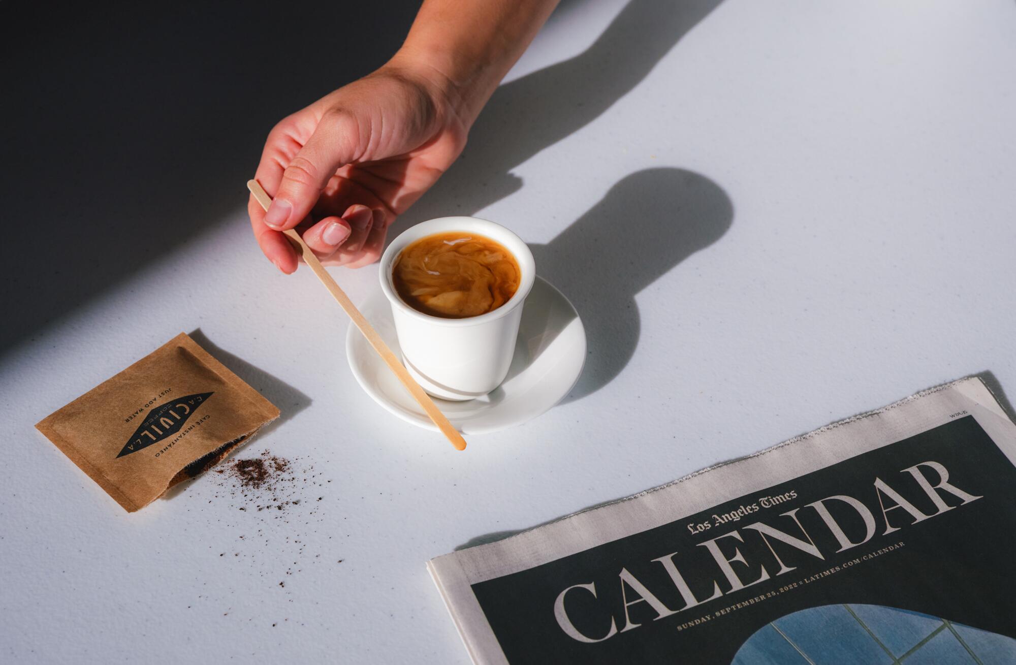 A hand holds a coffee stirrer next to a cup of coffee on a saucer, with an open packet of instant coffee next to it.
