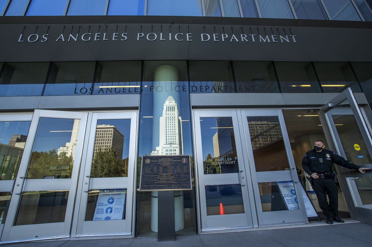 A LAPD officer wearing a face mask leaves police headquarters, where City Hall is reflected in a window.