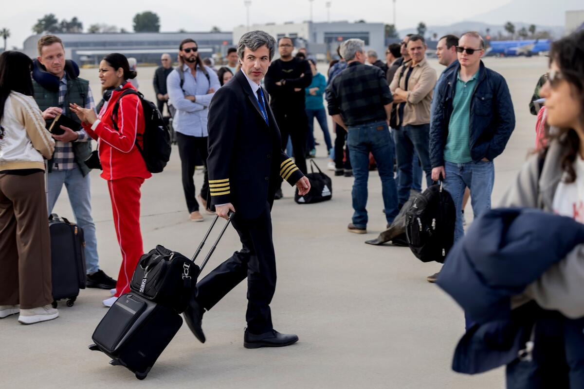 A man pulling luggage and dressed as a pilot walks through a crowd on a tarmac