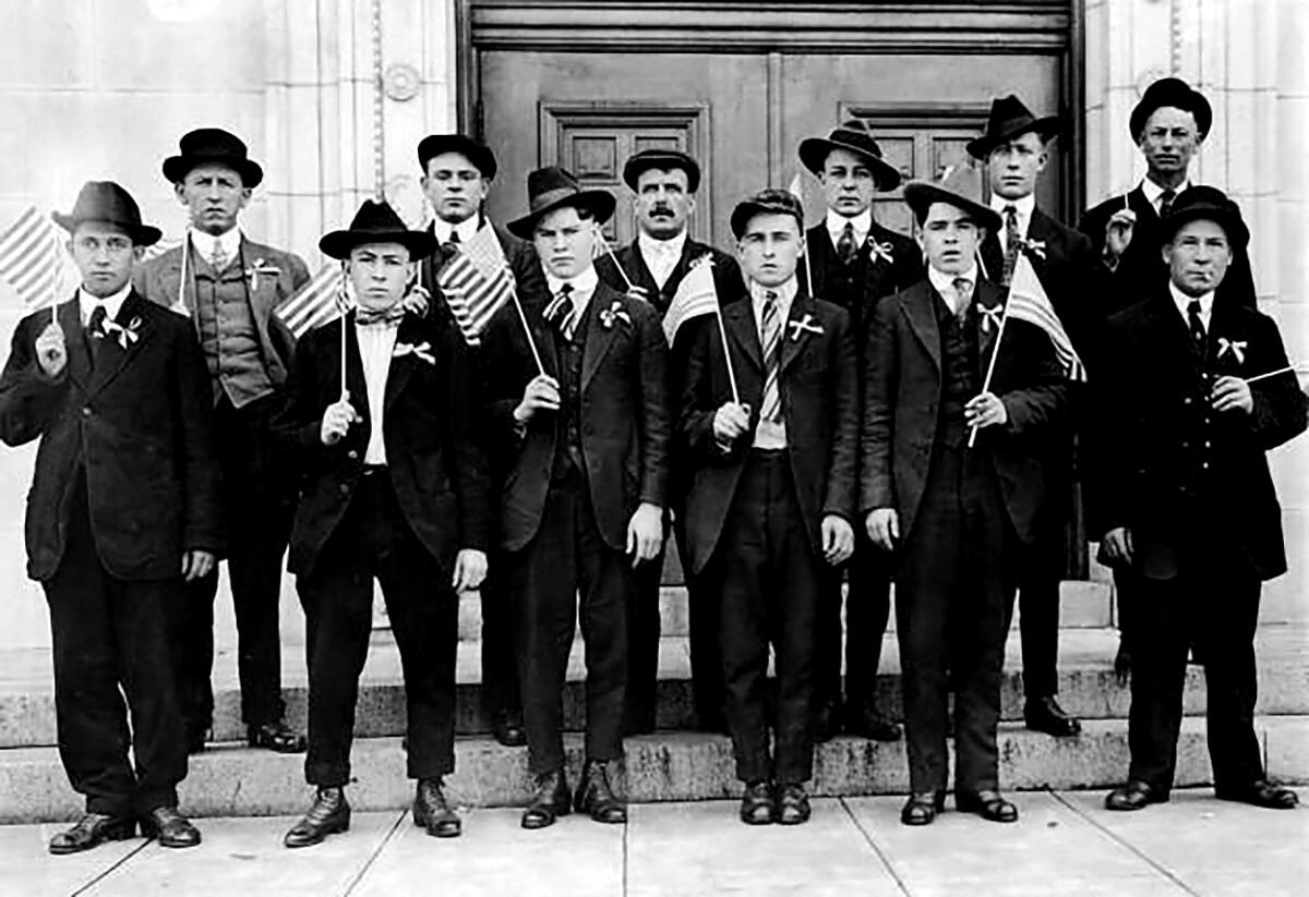 Men wearing 1910s suits and hats, holding flags, stand in two lines for a group portrait.