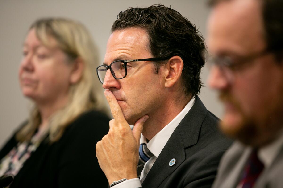 A man wearing a suit and glasses, with his hand to his face, sits between two other people listening intently.
