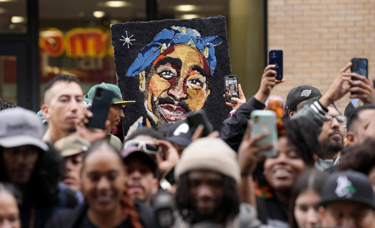 A painting of Tupac Shakur wearing a bandanna is held up among a crowd of people