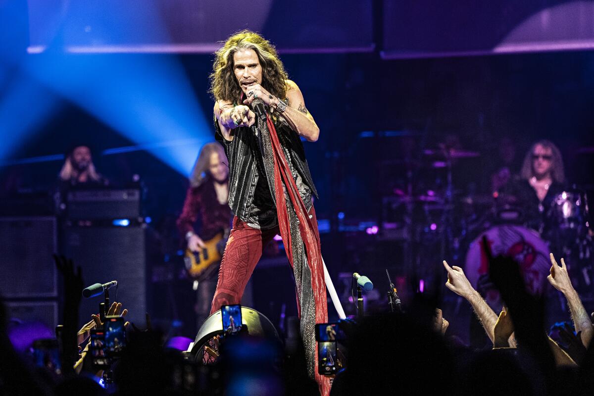 Steven Tyler wears long scarves draped around his neck and points toward the audience s he sings into a microphone