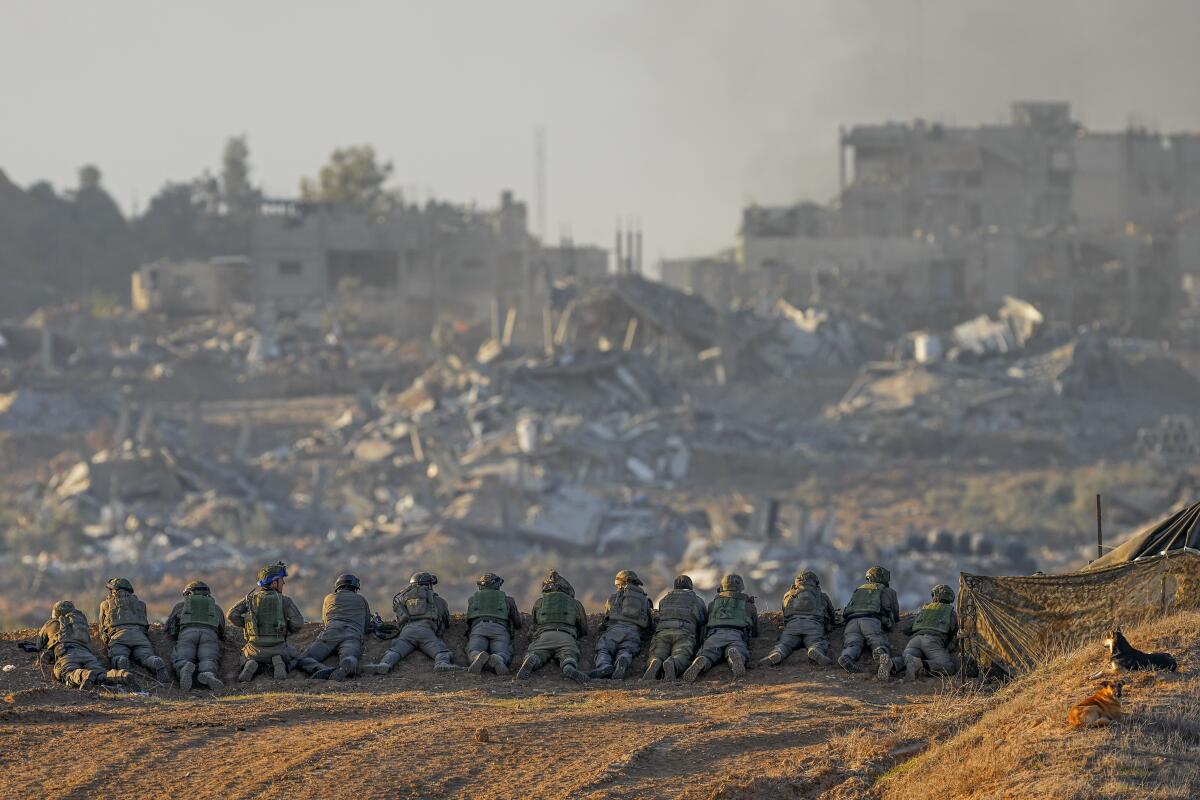 A line of soldiers in uniform are positioned at the edge of an overlook facing destroyed buildings 