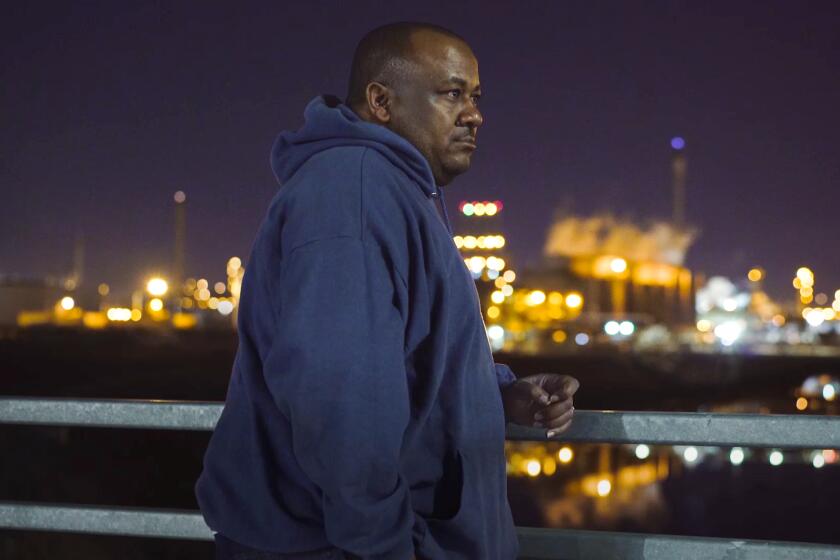 Image of a man in a navy hoodie standing outdoors at nighttime next to a railing with city lights behind him.
