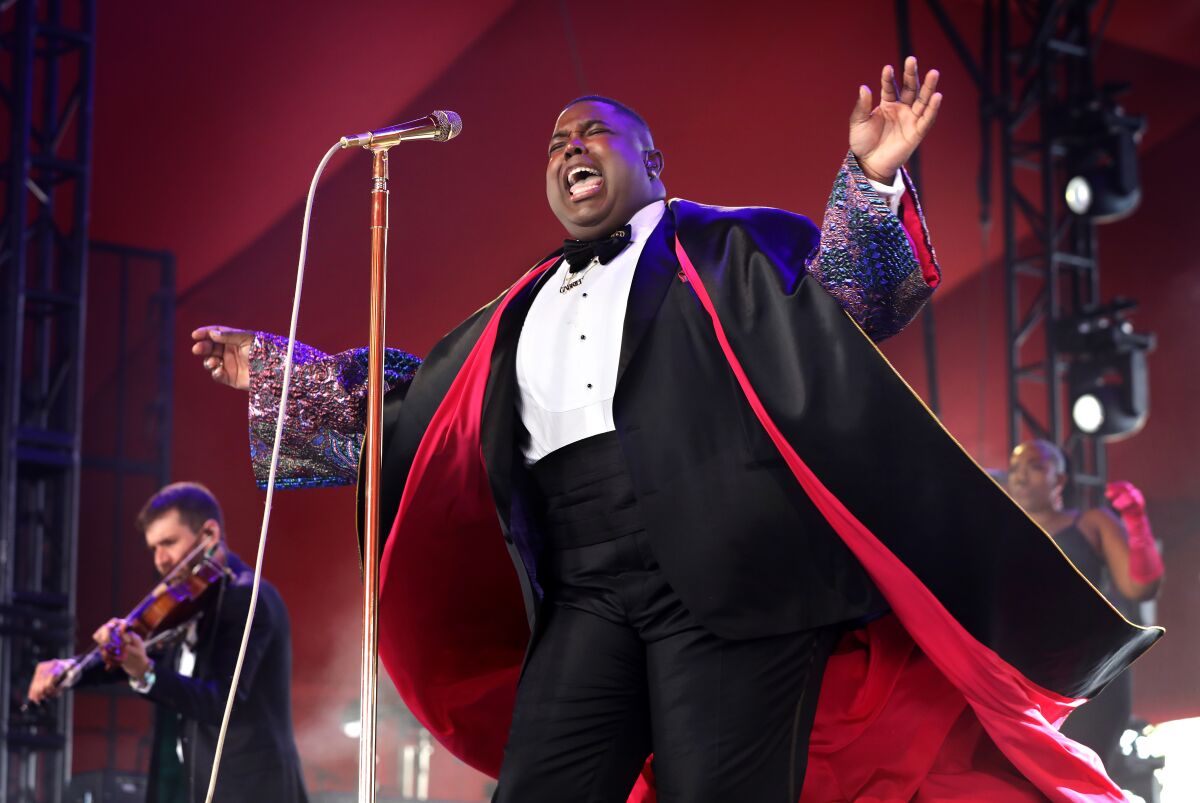 A man wearing a tuxedo sings on stage with his arms out to his sides