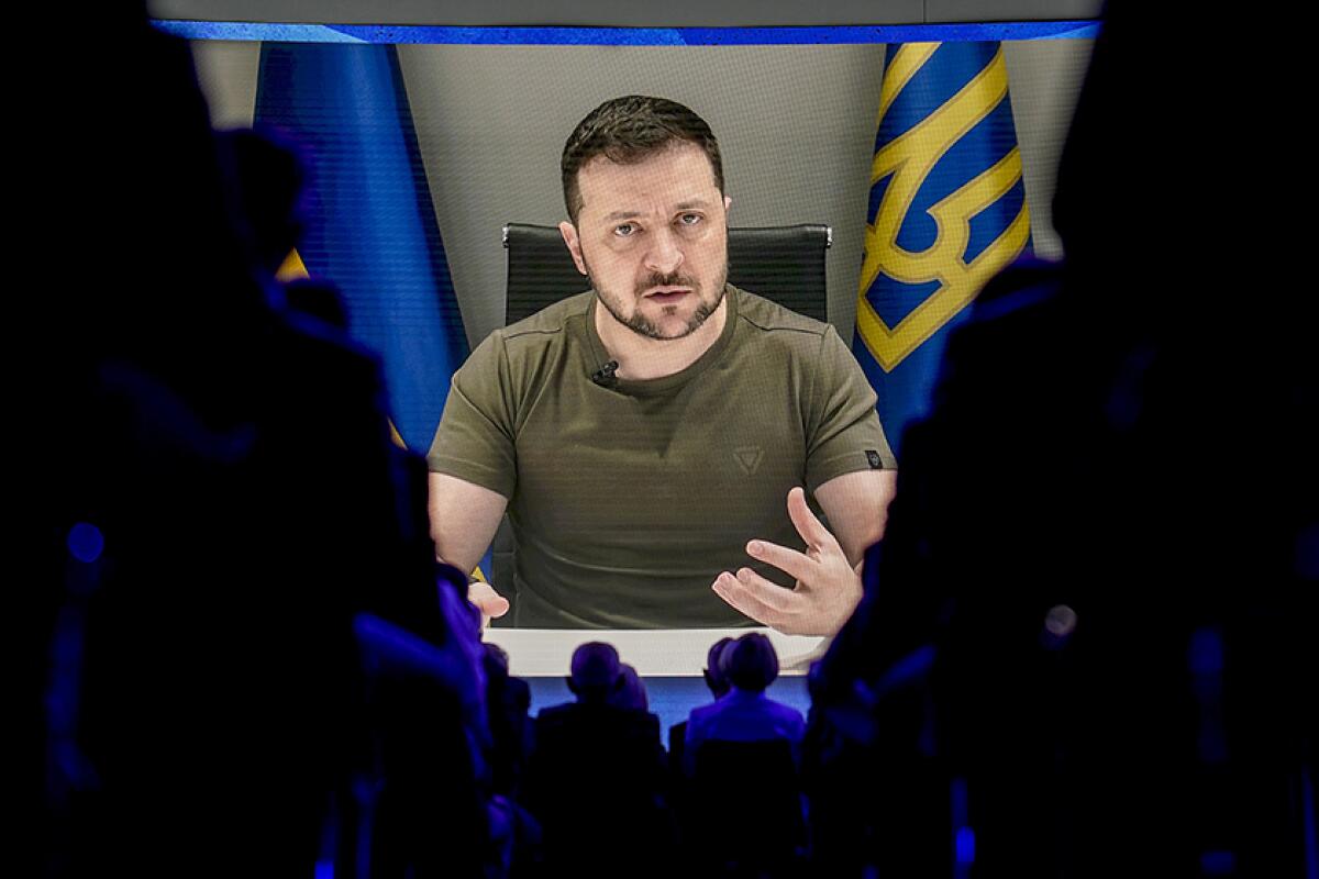 The image of Volodymyr Zelensky, speaking into microphones in front of flags, is projected on a screen 