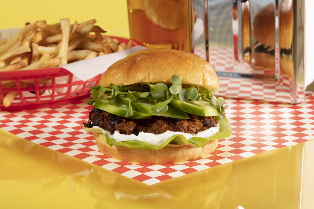 Close-up of a sweet potato burger wrapped in sesame, with fries and iced tea behind it, on a red and white checkered tablecloth