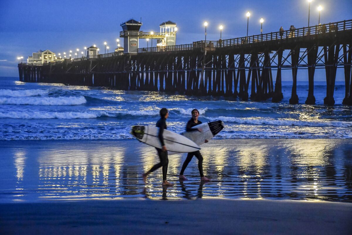 Two people carrying surfboards walk in the sand in front of a lighted pier in the evening.