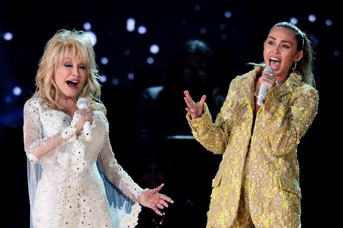 Dolly Parton, in a white dress with flower appliques, and Miley Cyrus, in a yellow and gold suit, perform onstage.